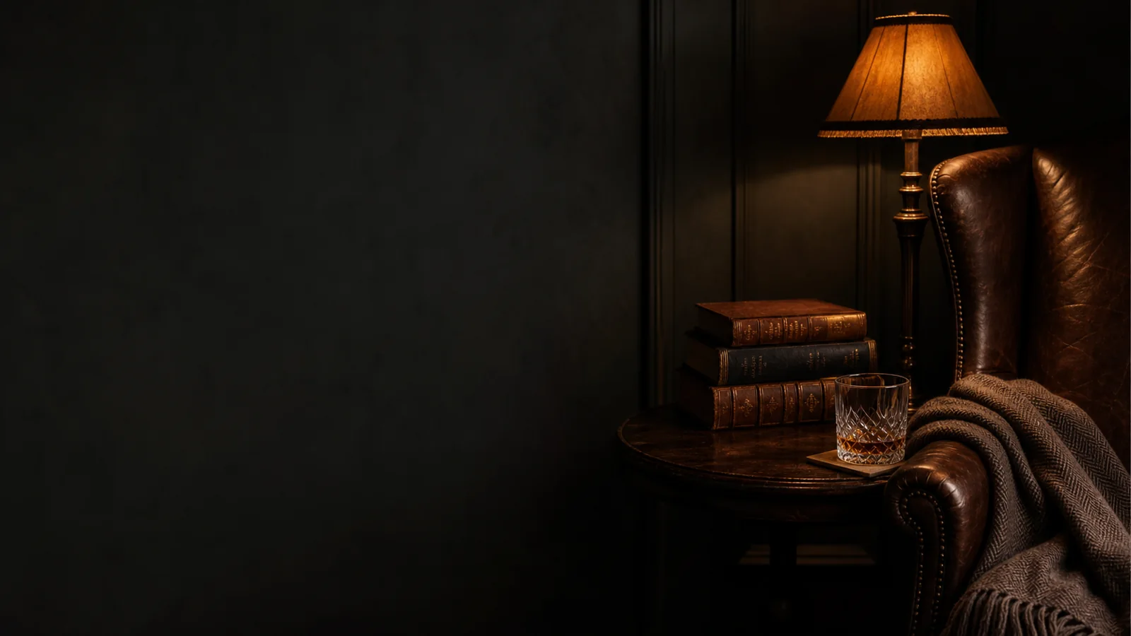 Editorial still-life of a leather wingback chair, stack of leather-bound books and a reading lamp in muted lamplight
